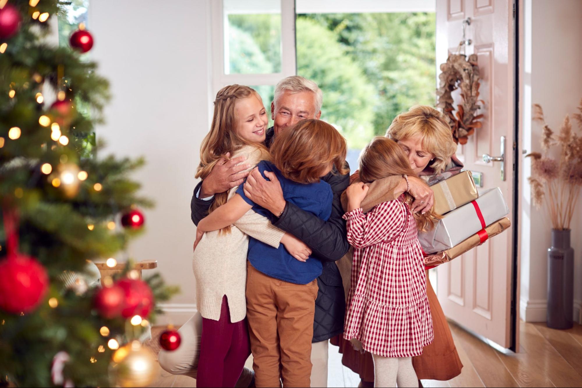 Family greeting grandchildren with holiday gifts in a warm, festive home. ©monkeybusiness